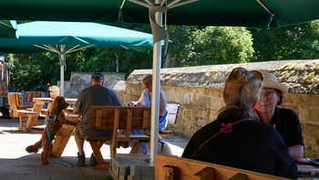 The photograph, taken in the morning during the summer, shows an outdoor restaurant area at Hardwick Hall in the United Kingdom. This National Trust property features wooden benches and tables under large green umbrellas, with groups of people seated and engaged in conversation. A dog is present, standing on its hind legs near one of the benches, and there is clear evidence that dogs are welcome at the restaurant. The scene is set against a stone wall with lush green trees in the background, representative of Hardwick Hall’s historic grounds. The image exemplifies an outdoor still life, capturing a moment of leisure among people enjoying the amenities of this notable National Trust landmark in the summer season.